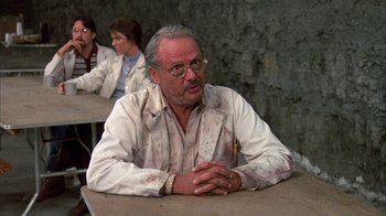 Movie still from “Day of the Dead” (1985), directed by George A. Romero – An older man sitting at a table in front of other people; Medium shot, Over the shoulder angle