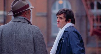 Movie still from “The Exorcist” (1973), directed by William Friedkin – A man wearing a coat and a hat standing next to another man; Medium shot, Over the shoulder angle