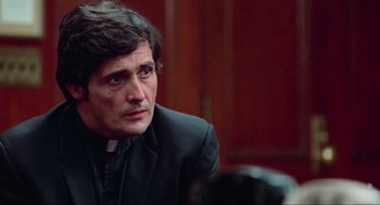 Movie still from “The Exorcist” (1973), directed by William Friedkin – A man wearing a priest's outfit sitting in front of a camera; Close Up shot, Over the shoulder angle
