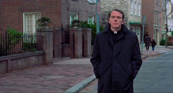 Movie still from “The Exorcist” (1973), directed by William Friedkin – A man standing on the side of the street wearing a black coat; Medium shot, Low angle