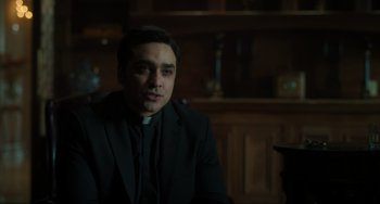 Movie still from “The Exorcist: Believer” (2023), directed by David Gordon Green – A man wearing a priest's outfit sitting in front of a bookshelf; Close Up shot, Low angle