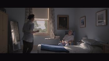 Movie still from “The Father” (2020), directed by Florian Zeller – An older man sitting in a chair next to a woman; Wide shot, High angle