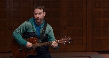 Movie still from “The Fault in Our Stars” (2014), directed by Josh Boone – A man is playing an acoustic guitar in front of a wall; Medium shot, Over the shoulder angle