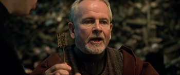 Movie still from “The Fifth Element” (1997), directed by Luc Besson – An older man with a beard holding a wooden object; Close Up shot, Over the shoulder angle