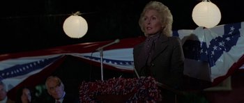Movie still from “The Fog” (1980), directed by John Carpenter – An older woman standing at a podium in front of an american flag; Medium shot, Low angle