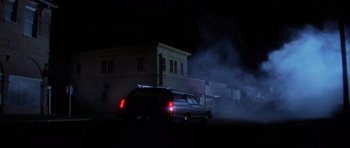 Movie still from “The Fog” (1980), directed by John Carpenter – A car driving down a street at night with smoke billowing from it; Extreme Wide shot, Low angle