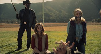 Movie still from “The Forever Purge” (2021), directed by Everardo Gout – A group of people standing on top of a grass covered field; Medium shot, Over the shoulder angle