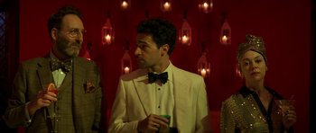 Movie still from “The Forgiven” (2021), directed by John Michael McDonagh – A man in a white suit and bow tie holding a drink; Medium shot, Over the shoulder angle