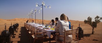 Movie still from “The Forgiven” (2021), directed by John Michael McDonagh – A group of people sitting at a long table in the middle of the desert; Wide shot, High angle
