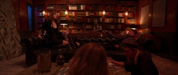 Movie still from “The Forgiven” (2021), directed by John Michael McDonagh – A group of people sitting on a black leather couch in a library; Wide shot, Low angle