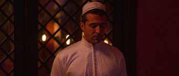 Movie still from “The Forgiven” (2021), directed by John Michael McDonagh – A man wearing a white hat standing in front of a fireplace; Medium shot, Over the shoulder angle