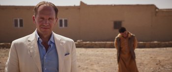 Movie still from “The Forgiven” (2021), directed by John Michael McDonagh – A man in a white suit standing next to a building; Medium shot, Over the shoulder angle