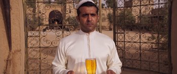 Movie still from “The Forgiven” (2021), directed by John Michael McDonagh – A man holding a glass of beer while wearing a white hat; Medium shot, Over the shoulder angle