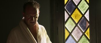 Movie still from “The Forgiven” (2021), directed by John Michael McDonagh – A man in a white bathrobe standing in front of a stained glass window; Close Up shot, Low angle