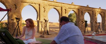 Movie still from “The Forgiven” (2021), directed by John Michael McDonagh – A man and a woman sitting in front of an arch bridge; Wide shot, Over the shoulder angle