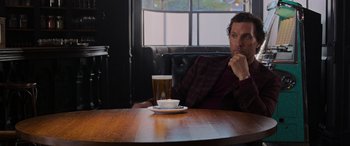 Movie still from “The Gentlemen” (2019), directed by Guy Ritchie – A man sitting at a table with a glass of beer in front of him; Medium shot, Over the shoulder angle