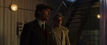 Movie still from “The Gentlemen” (2019), directed by Guy Ritchie – A man and a woman wearing hats standing next to each other; Medium shot, Low angle