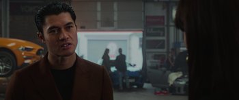 Movie still from “The Gentlemen” (2019), directed by Guy Ritchie – A man standing in front of a building with people in the background; Close Up shot, Over the shoulder angle