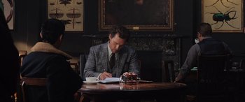 Movie still from “The Gentlemen” (2019), directed by Guy Ritchie – A man sitting at a table writing on a piece of paper; Medium shot, Over the shoulder angle