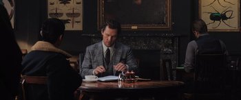 Movie still from “The Gentlemen” (2019), directed by Guy Ritchie – A man sitting at a wooden table in front of a book; Medium shot, Over the shoulder angle