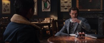 Movie still from “The Gentlemen” (2019), directed by Guy Ritchie – A man sitting at a table in front of another man drinking a cup; Medium shot, Over the shoulder angle