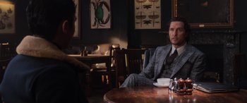 Movie still from “The Gentlemen” (2019), directed by Guy Ritchie – A man sitting at a table in a suit and tie; Medium shot, Over the shoulder angle