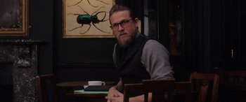 Movie still from “The Gentlemen” (2019), directed by Guy Ritchie – A man sitting at a wooden table in front of a painting; Medium shot, Over the shoulder angle