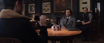 Movie still from “The Gentlemen” (2019), directed by Guy Ritchie – A man sitting at a wooden table in front of another man; Medium shot, Over the shoulder angle
