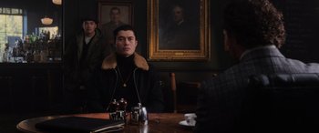 Movie still from “The Gentlemen” (2019), directed by Guy Ritchie – A man sitting at a table in front of a painting; Medium shot, Over the shoulder angle