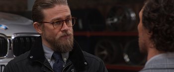 Movie still from “The Gentlemen” (2019), directed by Guy Ritchie – A man with a beard wearing glasses and a black jacket; Close Up shot, Over the shoulder angle