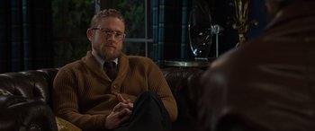 Movie still from “The Gentlemen” (2019), directed by Guy Ritchie – A man sitting on top of a couch in a living room; Close Up shot, Over the shoulder angle