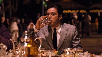 Movie still from “The Godfather Part II” (1974), directed by Francis Ford Coppola – A man in a gray suit drinking a glass of wine; Medium shot, Over the shoulder angle