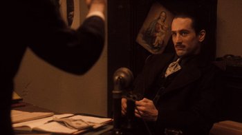 Movie still from “The Godfather Part II” (1974), directed by Francis Ford Coppola – A man sitting at a table in front of a microphone; Medium shot, Over the shoulder angle