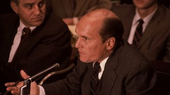 Movie still from “The Godfather Part II” (1974), directed by Francis Ford Coppola – A man in a suit and tie sitting in front of a microphone in front of other people; Close Up shot, Over the shoulder angle