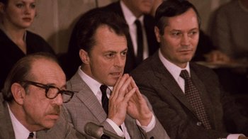 Movie still from “The Godfather Part II” (1974), directed by Francis Ford Coppola – A man in a gray suit clasps his hands while sitting in front of other men; Close Up shot, Over the shoulder angle