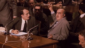 Movie still from “The Godfather Part II” (1974), directed by Francis Ford Coppola – Two men are sitting at a table in front of a microphone; Medium shot, Over the shoulder angle