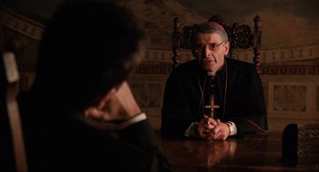 Movie still from “The Godfather Part III” (1990), directed by Francis Ford Coppola – A man sitting at a table holding a cross in front of another man; Medium shot, Over the shoulder angle