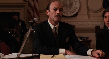 Movie still from “The Godfather Part III” (1990), directed by Francis Ford Coppola – A man sitting at a table in front of a microphone; Medium shot, Low angle