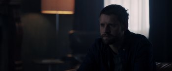 Movie still from “The Good Neighbor” (2022), directed by Stephan Rick – A man sitting at a table in a dark room; Close Up shot, Over the shoulder angle