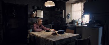 Movie still from “The Good Neighbor” (2022), directed by Stephan Rick – An older woman sitting at a table in a kitchen; Wide shot, High angle