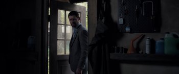 Movie still from “The Good Neighbor” (2022), directed by Stephan Rick – A man in a gray suit standing in front of an open door; Medium shot, Low angle