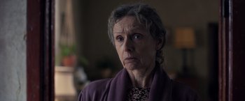Movie still from “The Good Neighbor” (2022), directed by Stephan Rick – An older woman wearing a purple jacket looking at the camera; Close Up shot, Over the shoulder angle