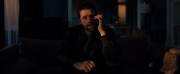 Movie still from “The Good Neighbor” (2022), directed by Stephan Rick – A man sitting on a couch holding a cell phone to his ear; Medium shot, Low angle