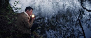 Movie still from “The Good Neighbor” (2022), directed by Stephan Rick – A man lighting a candle in front of a body of water; Medium shot, High angle