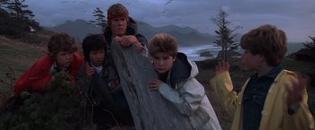 Movie still from “The Goonies” (1985), directed by Richard Donner – A group of young people standing next to each other on a beach; Medium shot, Low angle