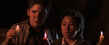 Movie still from “The Goonies” (1985), directed by Richard Donner – Two young men holding a lit candle in a dark room; Close Up shot, Low angle