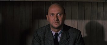 Movie still from “The Great Escape” (1963), directed by John Sturges – A man in a suit and tie sitting in front of a wooden wall; Close Up shot, Low angle