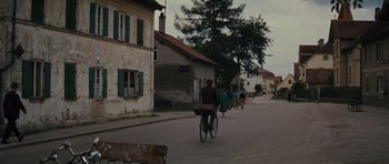 Movie still from “The Great Escape” (1963), directed by John Sturges – A man riding a bike down a street with people; Extreme Wide shot, Low angle