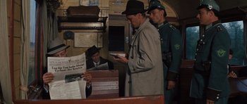 Movie still from “The Great Escape” (1963), directed by John Sturges – A man in a hat and a coat is looking at a newspaper; Medium shot, Over the shoulder angle