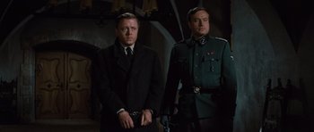 Movie still from “The Great Escape” (1963), directed by John Sturges – A man in a suit standing next to another man in a uniform; Medium shot, Low angle